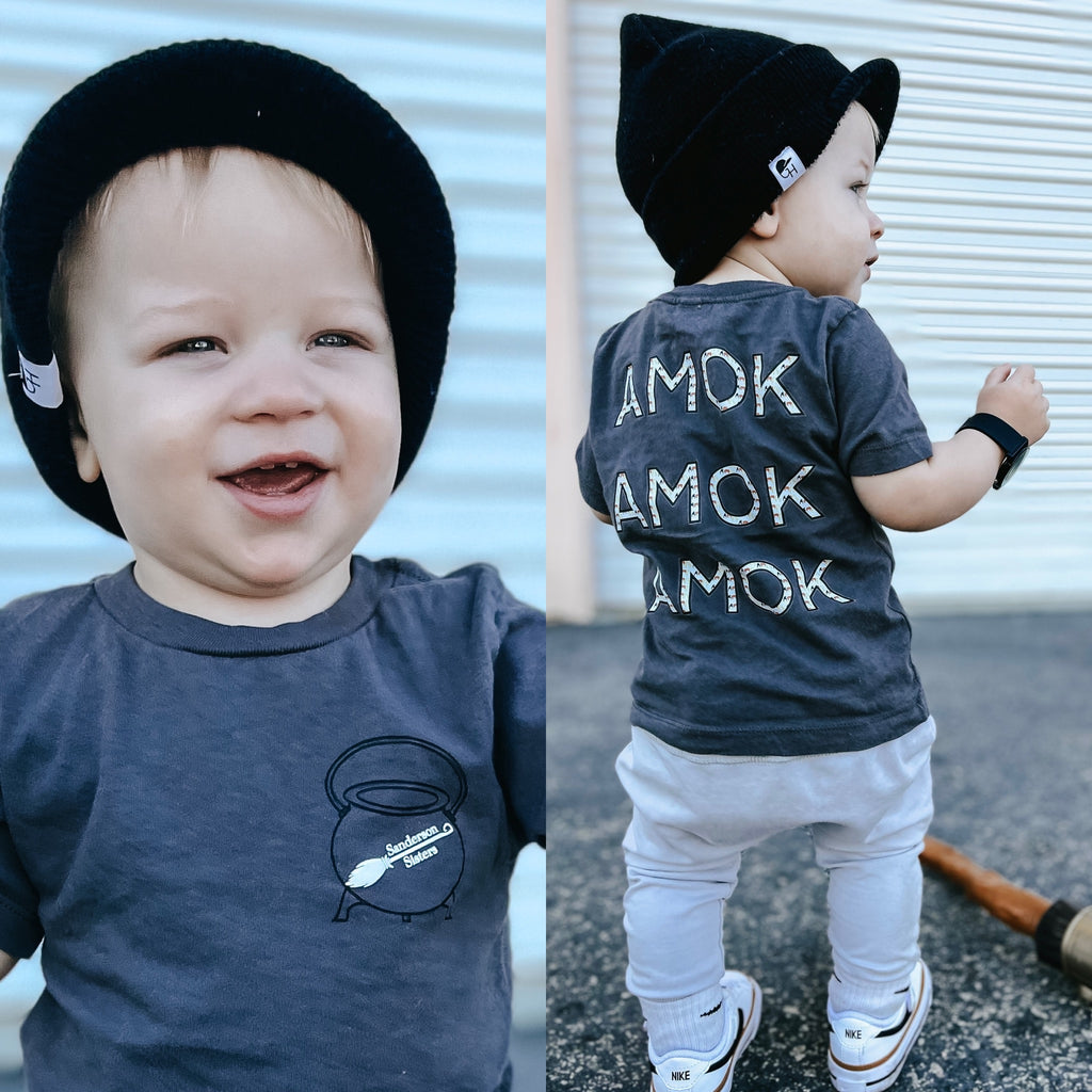 1 year old boy wearing a Hocus Pocus shirt. The shirt had a cauldron on the front with a white witches broom with the words Sanderson Sisters. The back has the words Amok Amok Amok centered with the silhouette of the Sanderson Sisters hair filled inside the letters.