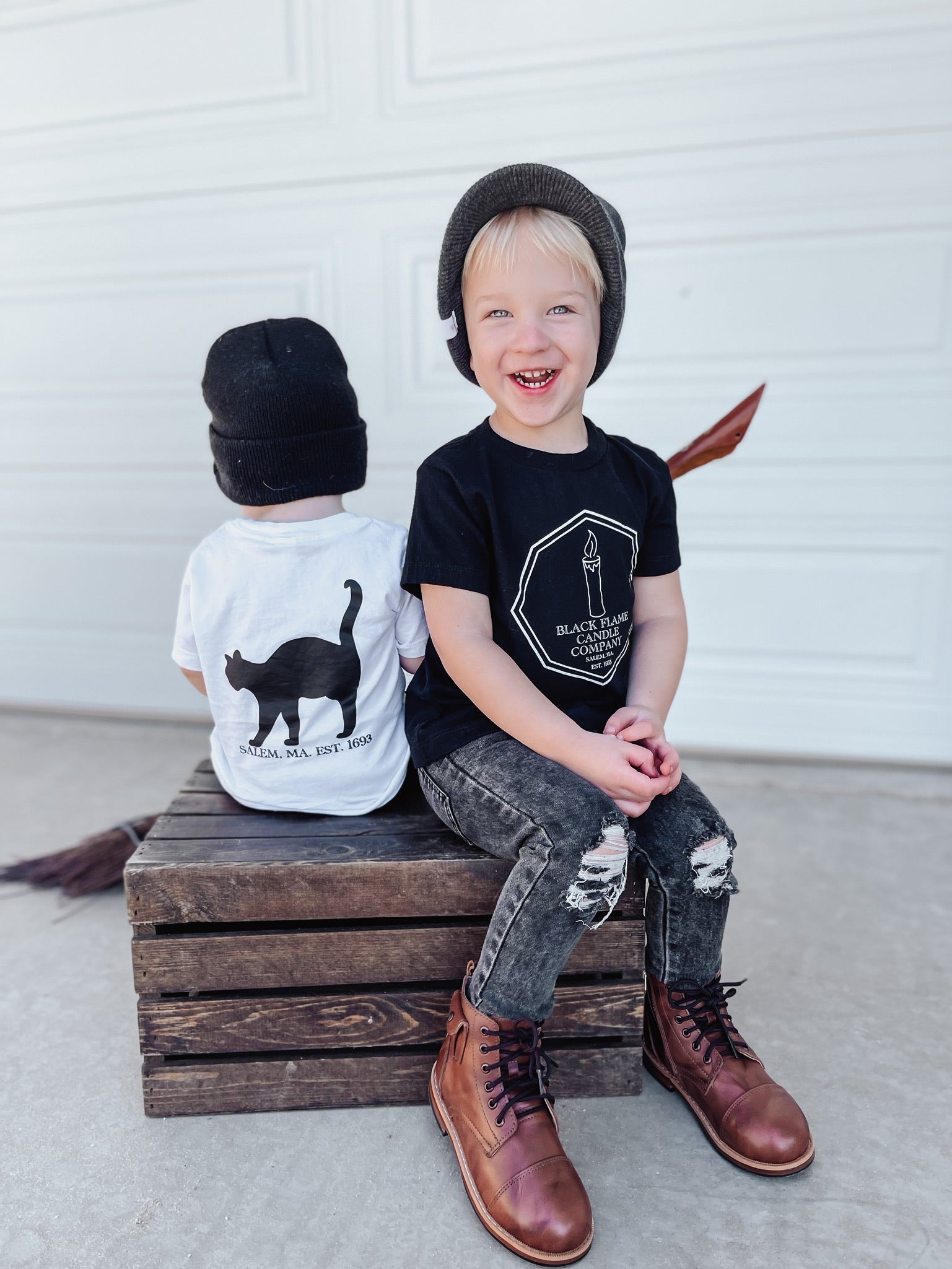2 little boys wearing Hocus Pocus shirts. The shirt has a "Binx Candle Security" badge on the front left breast pocket area. The back of the shirt has a silhouette of a black cat and the words Salem, MA. EST. 1693 under it. 