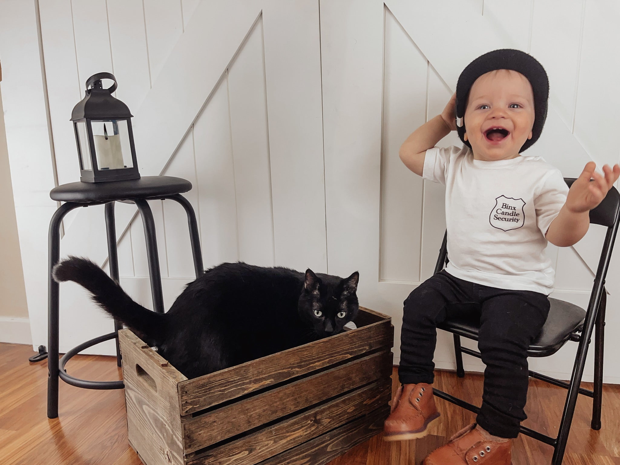 1 year old boy wearing a Hocus Pocus shirt. The shirt has a "Binx Candle Security" badge on the front left breast pocket area. The back of the shirt has a silhouette of a black cat and the words Salem, MA. EST. 1693 under it. There is a black cat crouched down in the photo next to the child. 